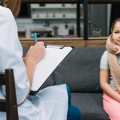 female-doctor-writing-clipboard-with-pen-front-girl-sitting-sofa-min